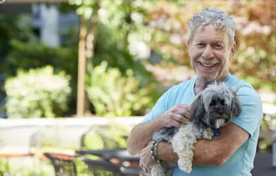 Happy male resident outdoors, holding a small white dog, Viamonte at Walnut Creek Lon 250th anniversary