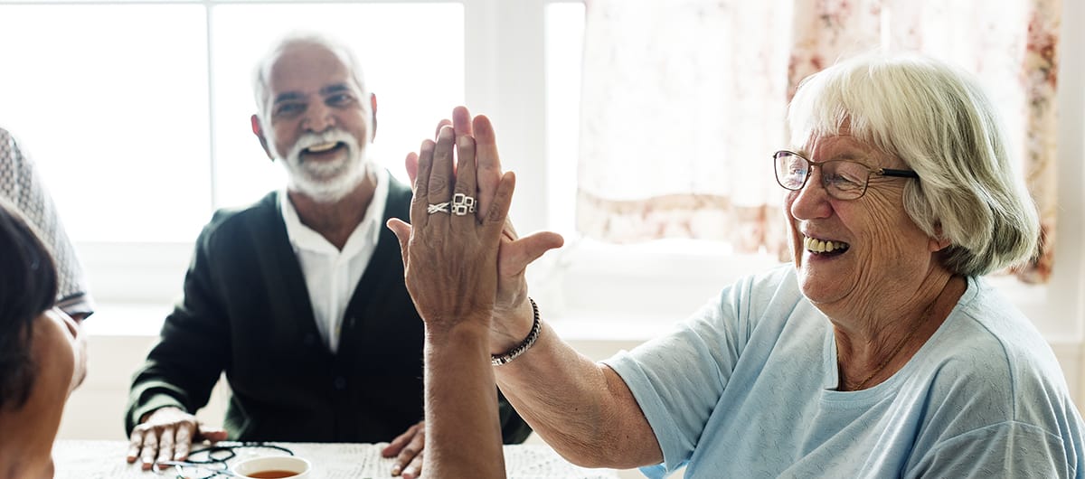 Assisted Living and Memory Care. Two elderly women high-fiving. Elderly man in background