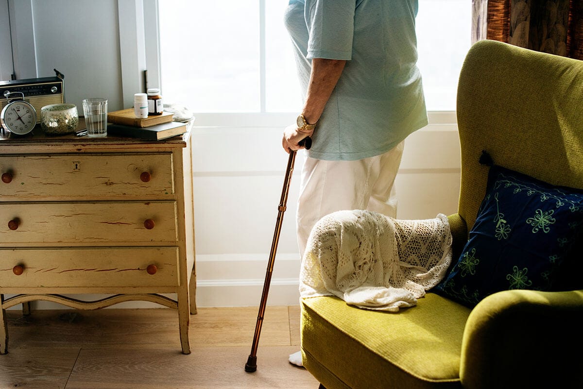Elderly woman standing next to green chair, holding a cane.