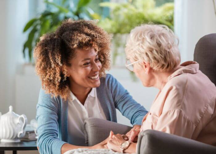 Image shows younger woman, a nurse or caregiver, sitting and talking with an older senior woman