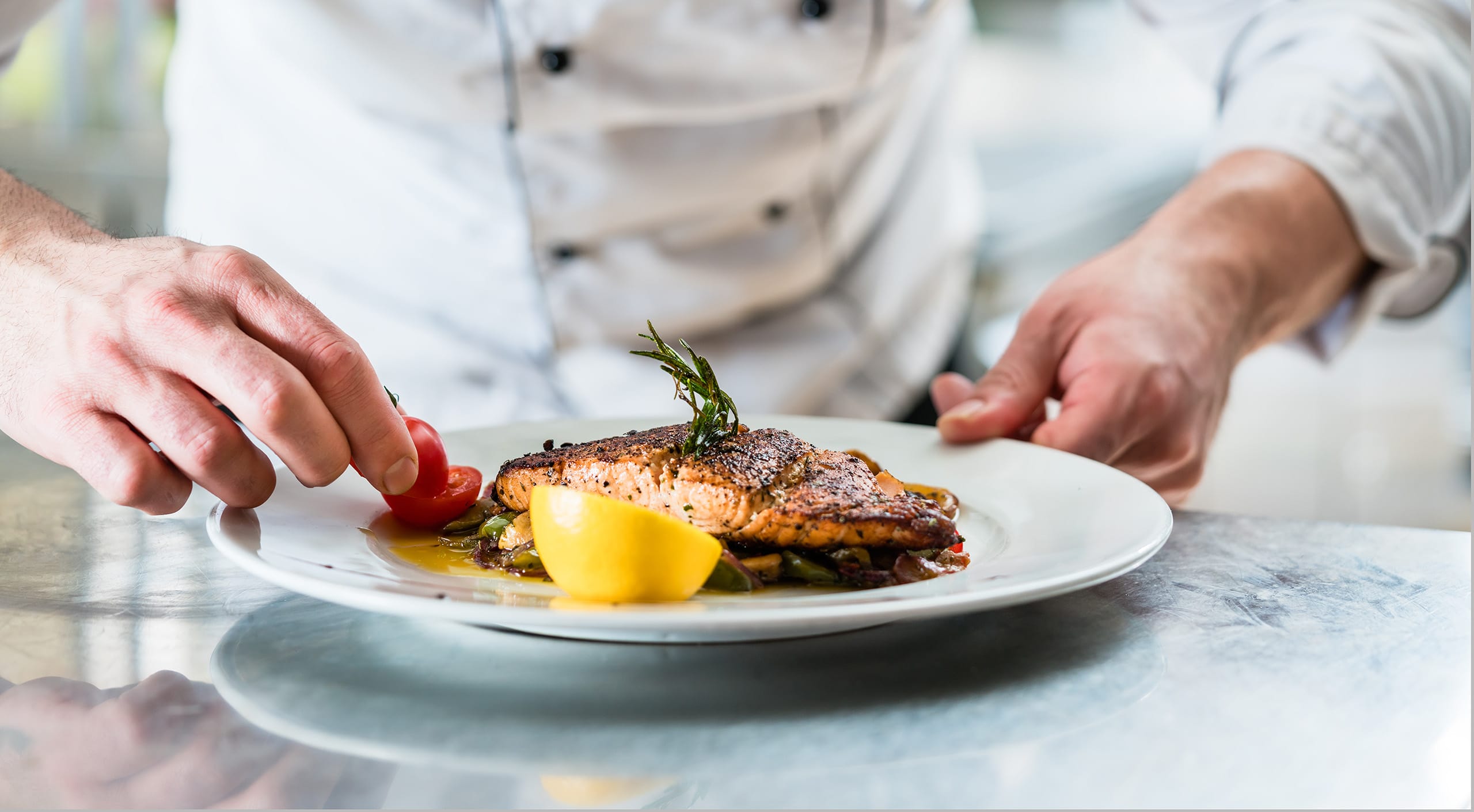 A chef's hands prepare a plate with grilled salmon, tomatoes and lemon to show healthy dining options
