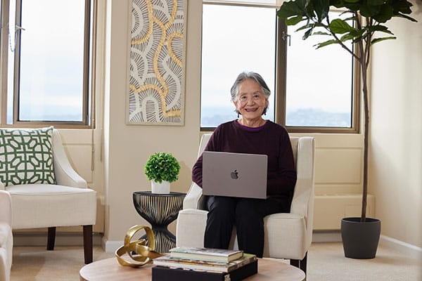 Older adult woman sits in apartment with a laptop computer, smiling at the camera