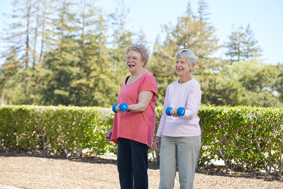 Two female residents wearing pink workout tops stand lifting single 4 pound weights and smiling at the camera