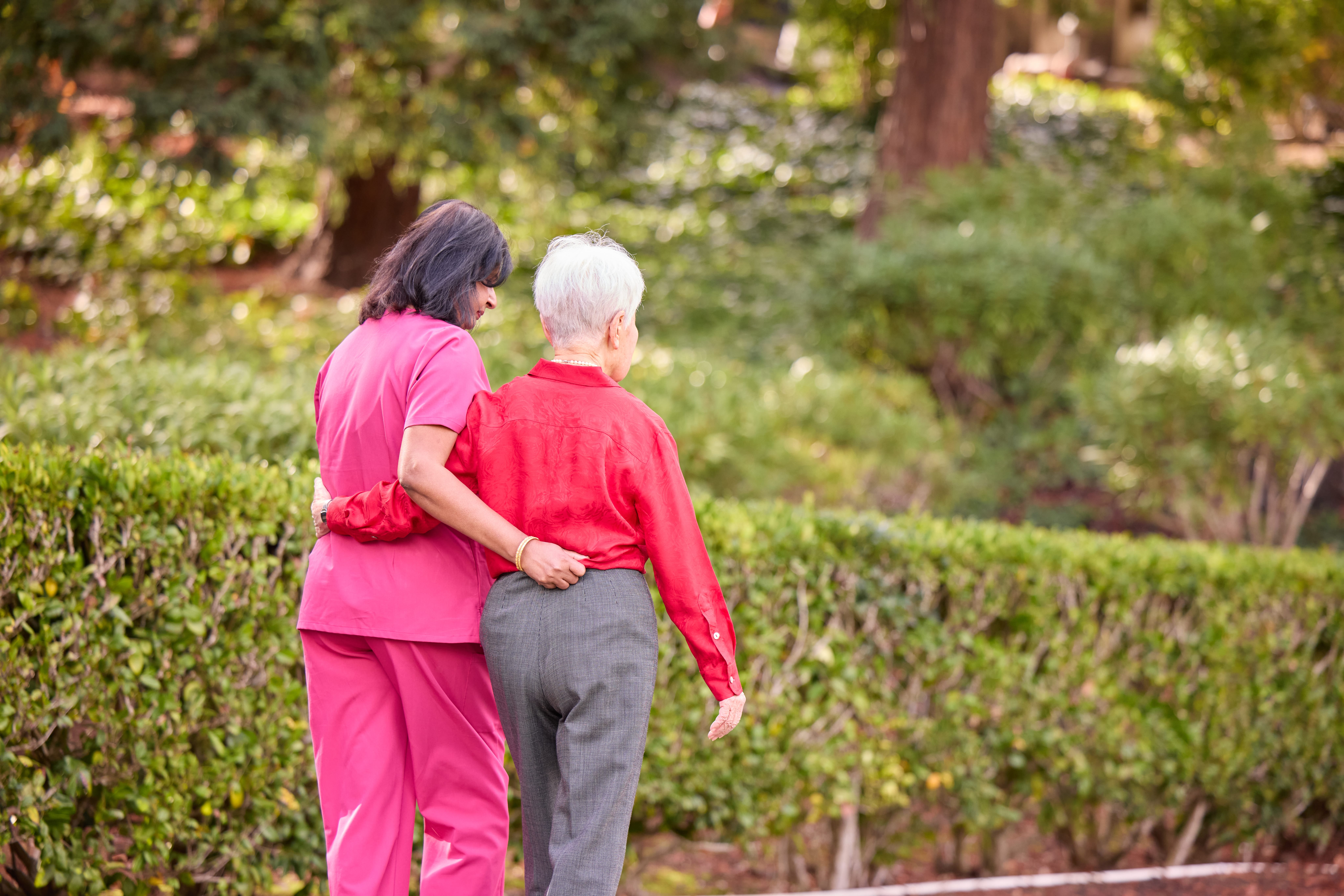 Elderly lady and nurse walking next to green shrubs