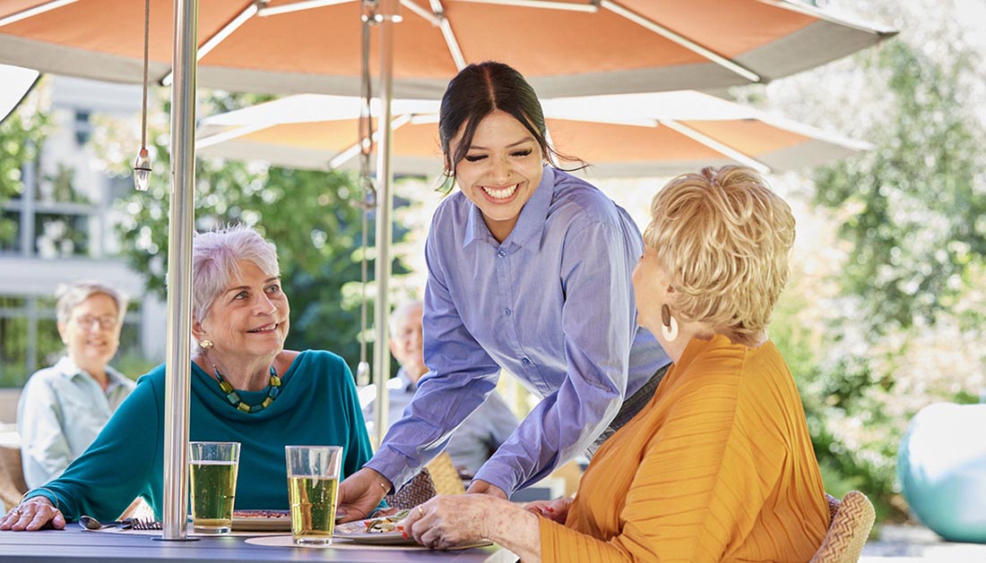 Enhanced dining experience | a dining staff person serving food to two older adult women, residents